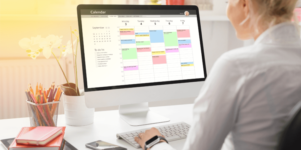 A woman sitting in front of a computer planning a schedule with some books and a phone nearby.