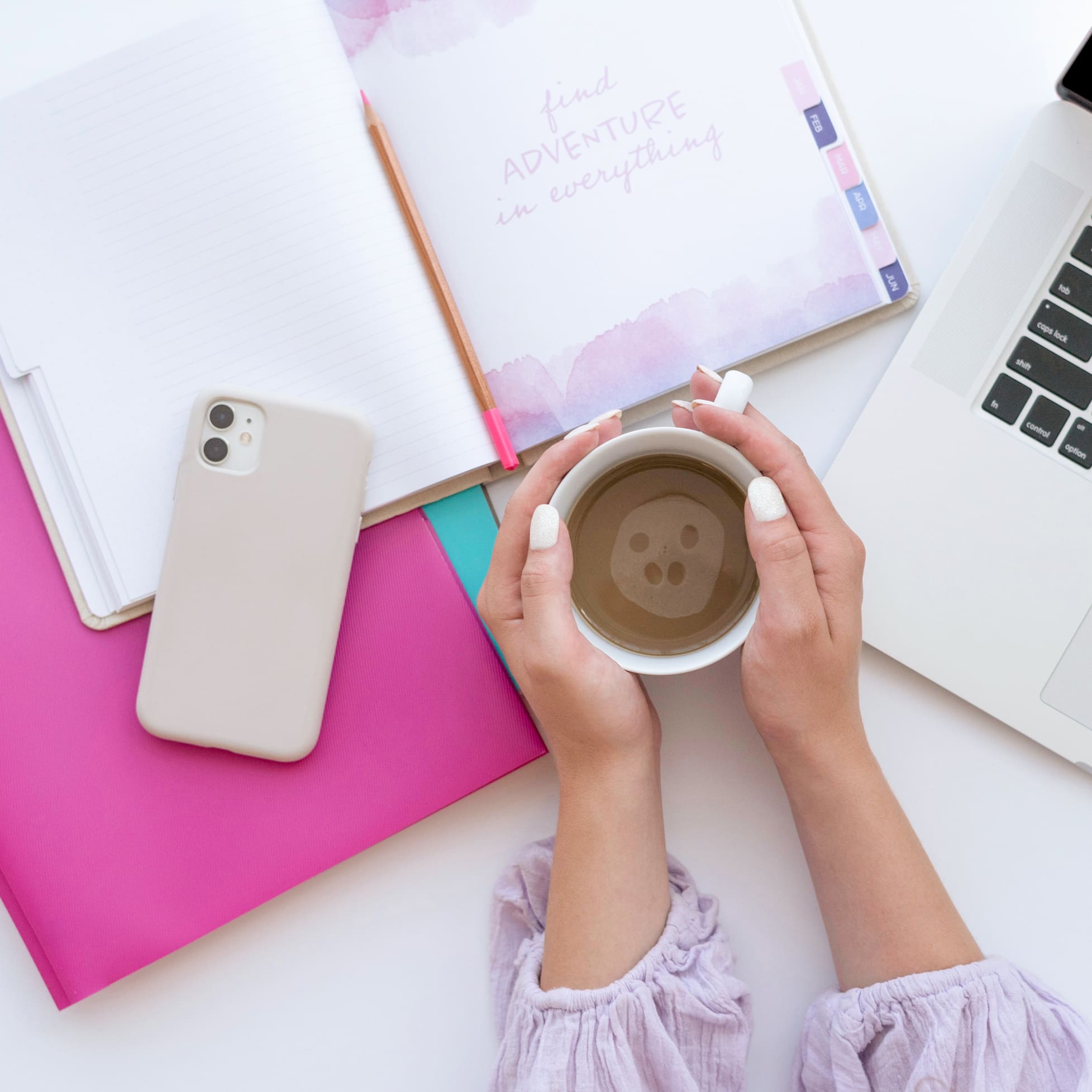 picture of a woman holding a cup of coffee between both hands on a table with a laptop, cell phone, folders, and a planner open with the words "find adventure in everything" written on the open page.